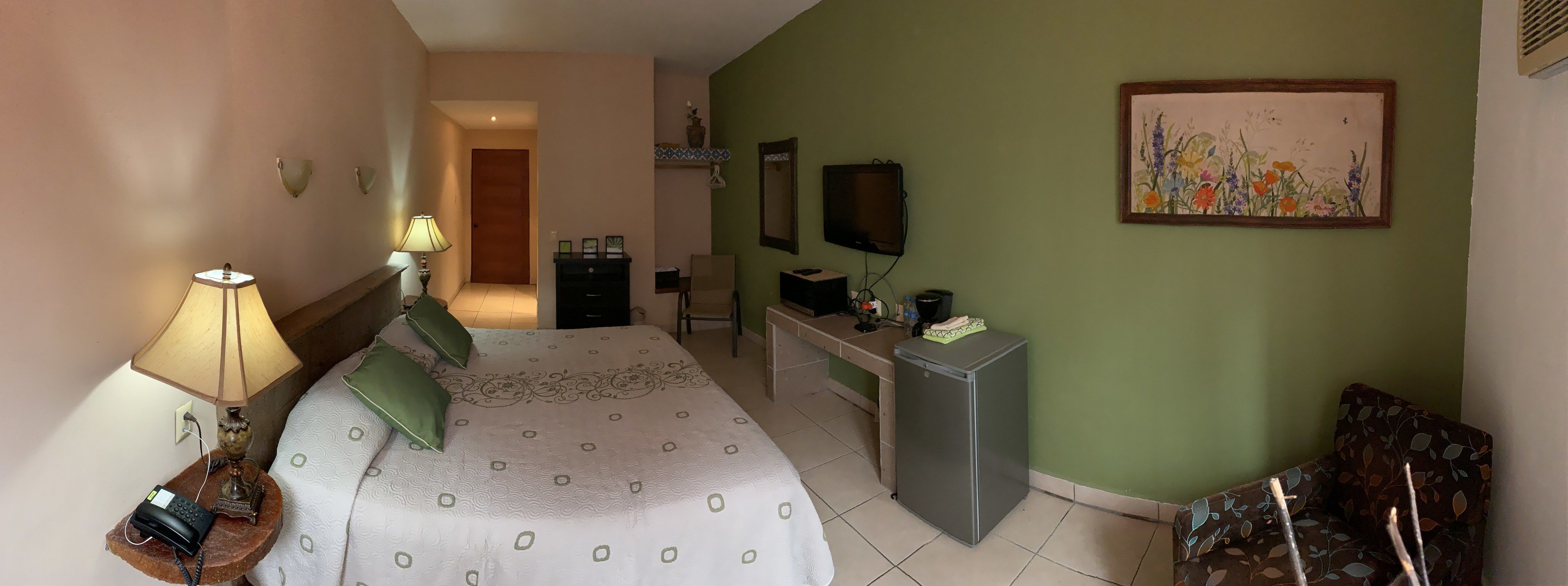 A hotel room with a bed, bedside lamp, desk, TV, and a painting on the wall.