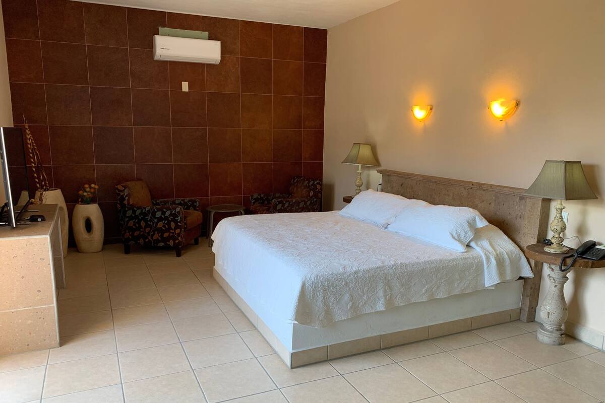 Une chambre d’hôtel avec un grand lit, deux lampes de chevet, un bureau avec une lampe, une chaise et un climatiseur fixé au mur.