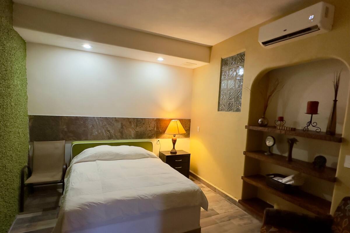 Une chambre d’hôtel comprenant un lit, une chaise, une table de chevet avec une lampe, un appareil de climatisation et une étagère avec des objets décoratifs.