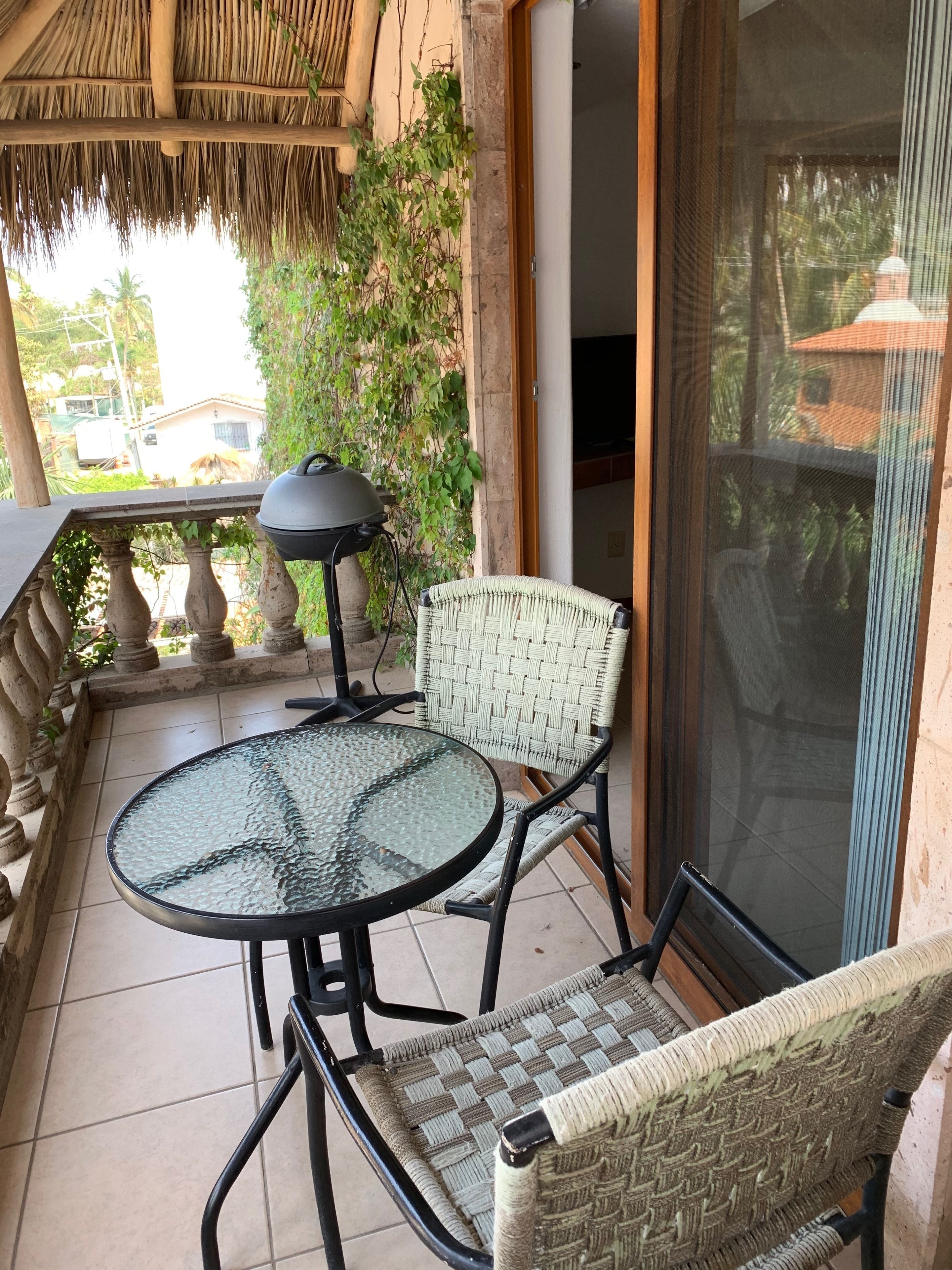 A balcony with a glass table and wicker chairs, a grill, and a view of a house and palm trees.