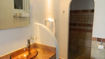 Une salle de bain avec un lavabo en verre, un miroir, un porte-serviettes, une douche aux murs carrelés et des toilettes.