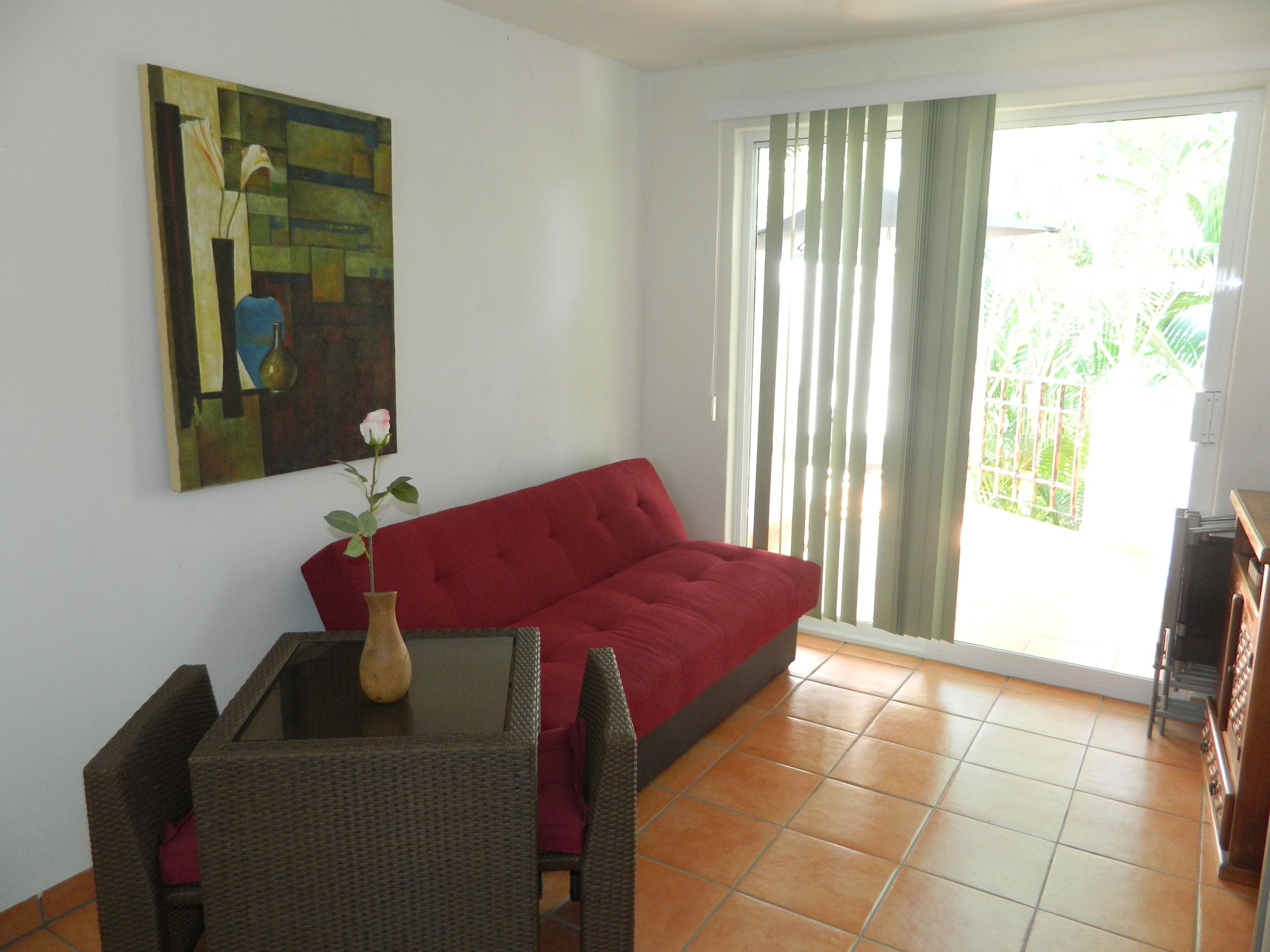 Una sala de estar con un sofá rojo, una mesa de vidrio, un florero con una flor y un cuadro en la pared.