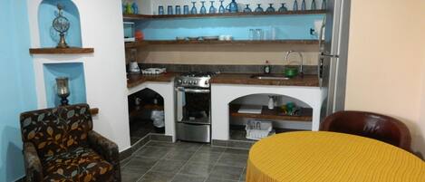 A compact kitchen with a stove, oven, and shelves above the counter. There is a dining table with a yellow tablecloth and a patterned armchair.