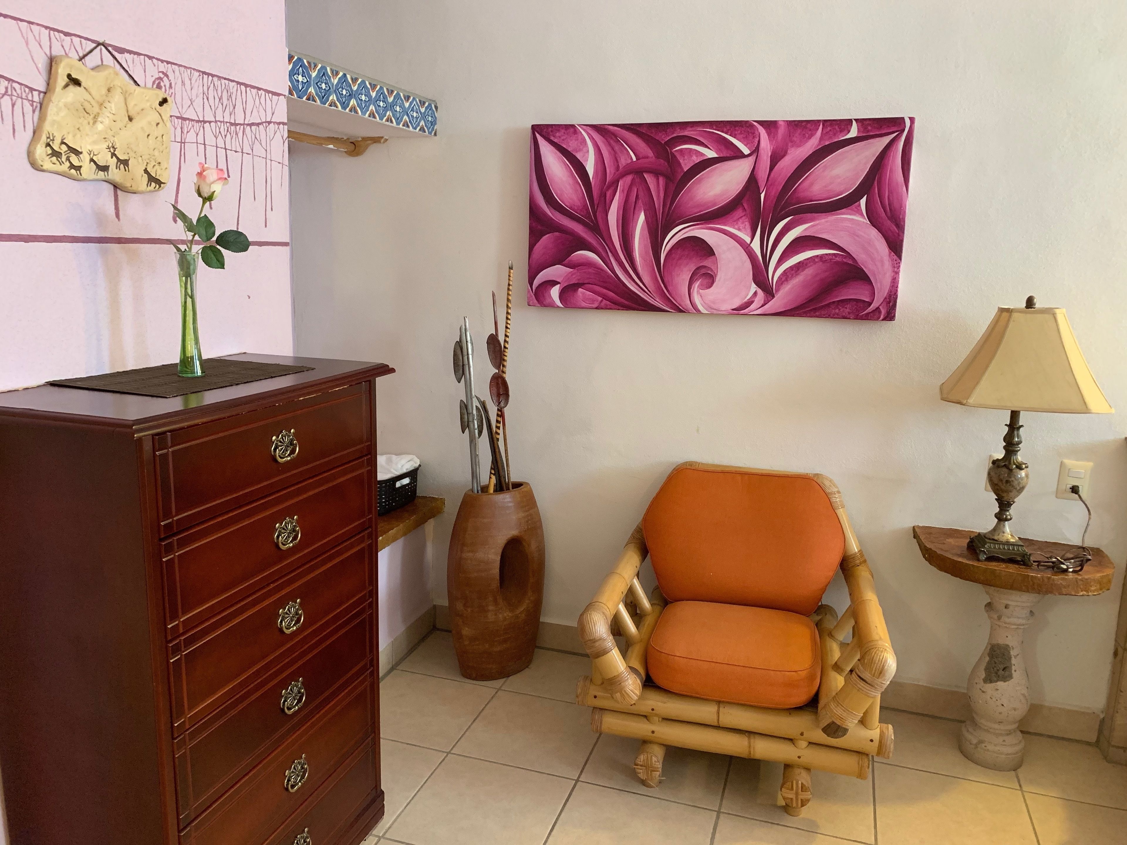 Une pièce comprenant une commode en bois, une chaise, un vase avec une fleur, un tableau et une lampe.