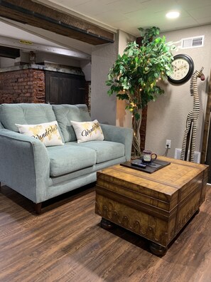 A living room with a light blue sofa, two pillows with 'Wanderlust' text, a wooden coffee table, a large plant, a clock, and a giraffe decoration.