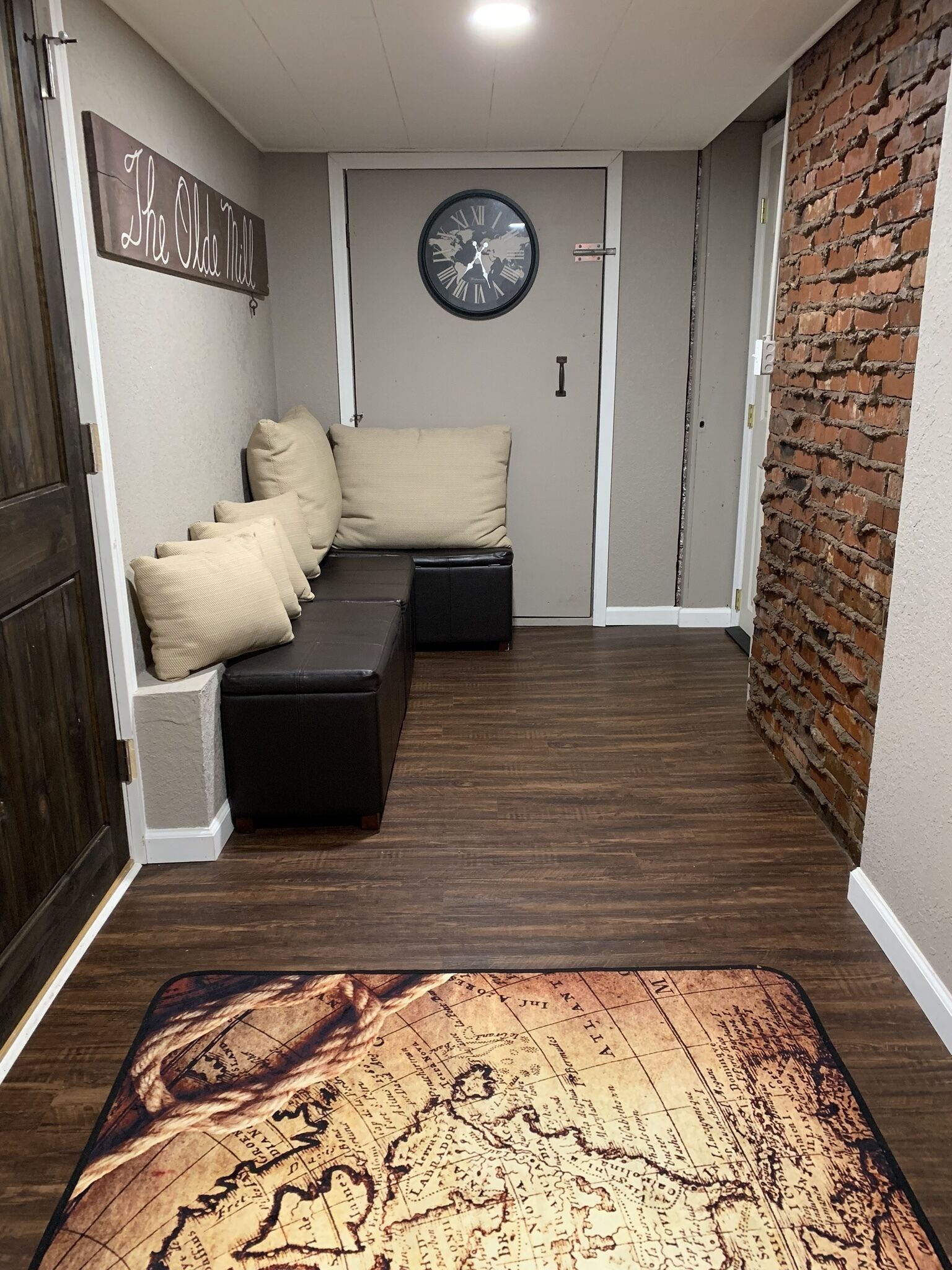 A hallway with a dark wood floor, a black leather bench with beige cushions, a world map clock, and a brick wall.
