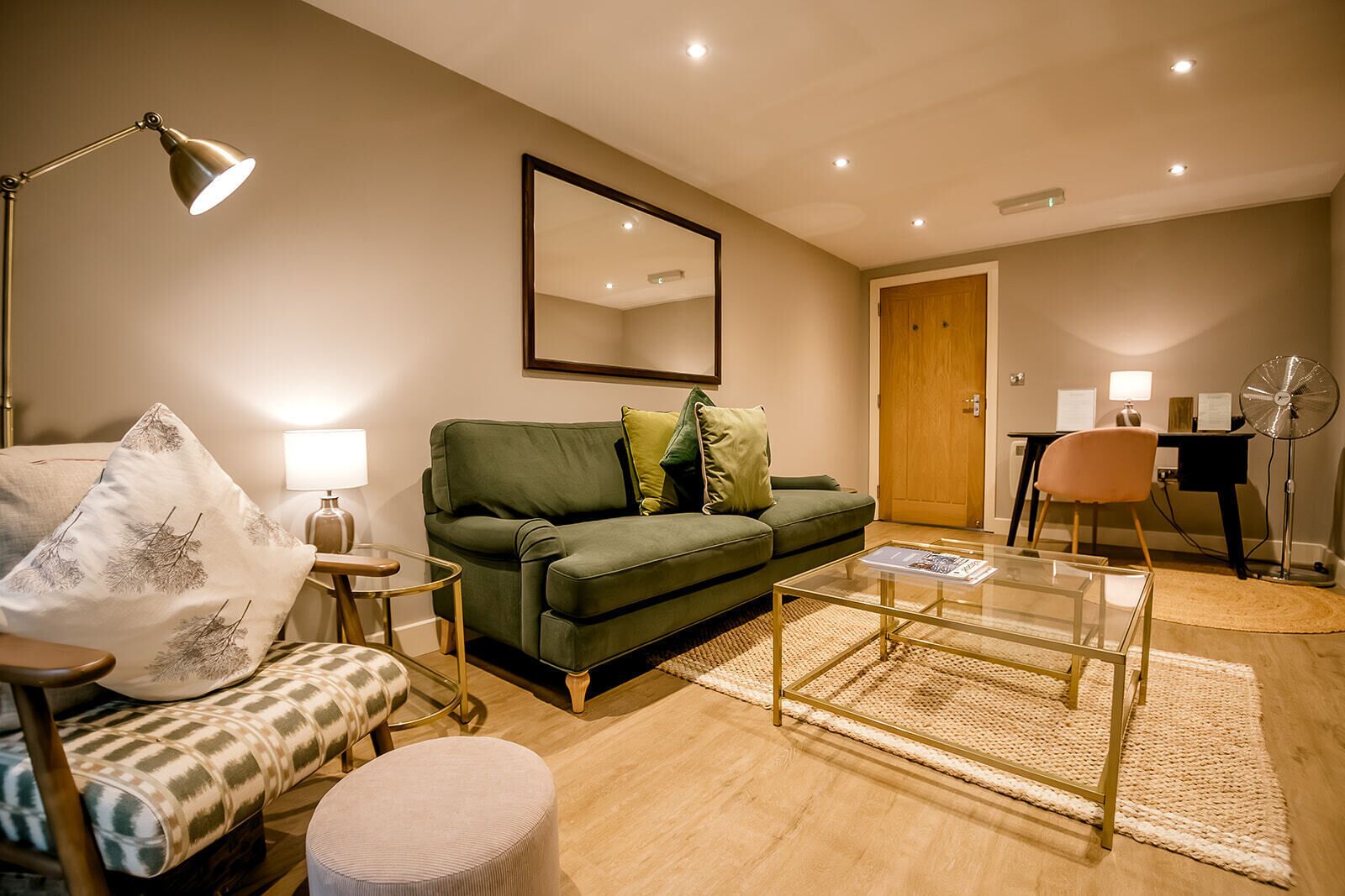 A living room with a green sofa, a small round ottoman, a glass coffee table, a floor lamp, a chair with a patterned cushion, and a mirror on the wall.