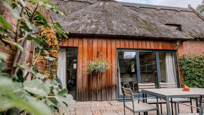 A rustic building with a thatched roof, wooden walls, and a large sliding glass door leading to an outdoor seating area with tables and chairs. - Norfolk Mead (Norwich)
