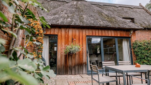 Il s’agit d’un bâtiment rustique doté d’un toit de chaume, de murs en bois et d’une grande porte-fenêtre coulissante qui donne sur un espace extérieur aménagé avec des tables et des chaises.