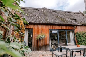 A rustic building with a thatched roof, wooden walls, and a large sliding glass door leading to an outdoor seating area with tables and chairs. - Norfolk Mead (Norwich)