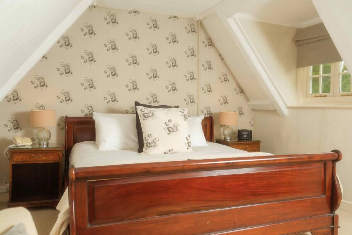 A bedroom with a wooden bed, two bedside tables with lamps, a patterned wallpaper, and a window with a view.