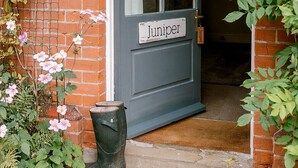Une porte portant l’inscription « Juniper », un mur de briques, une fenêtre avec un rideau et une botte verte posée sur le seuil.