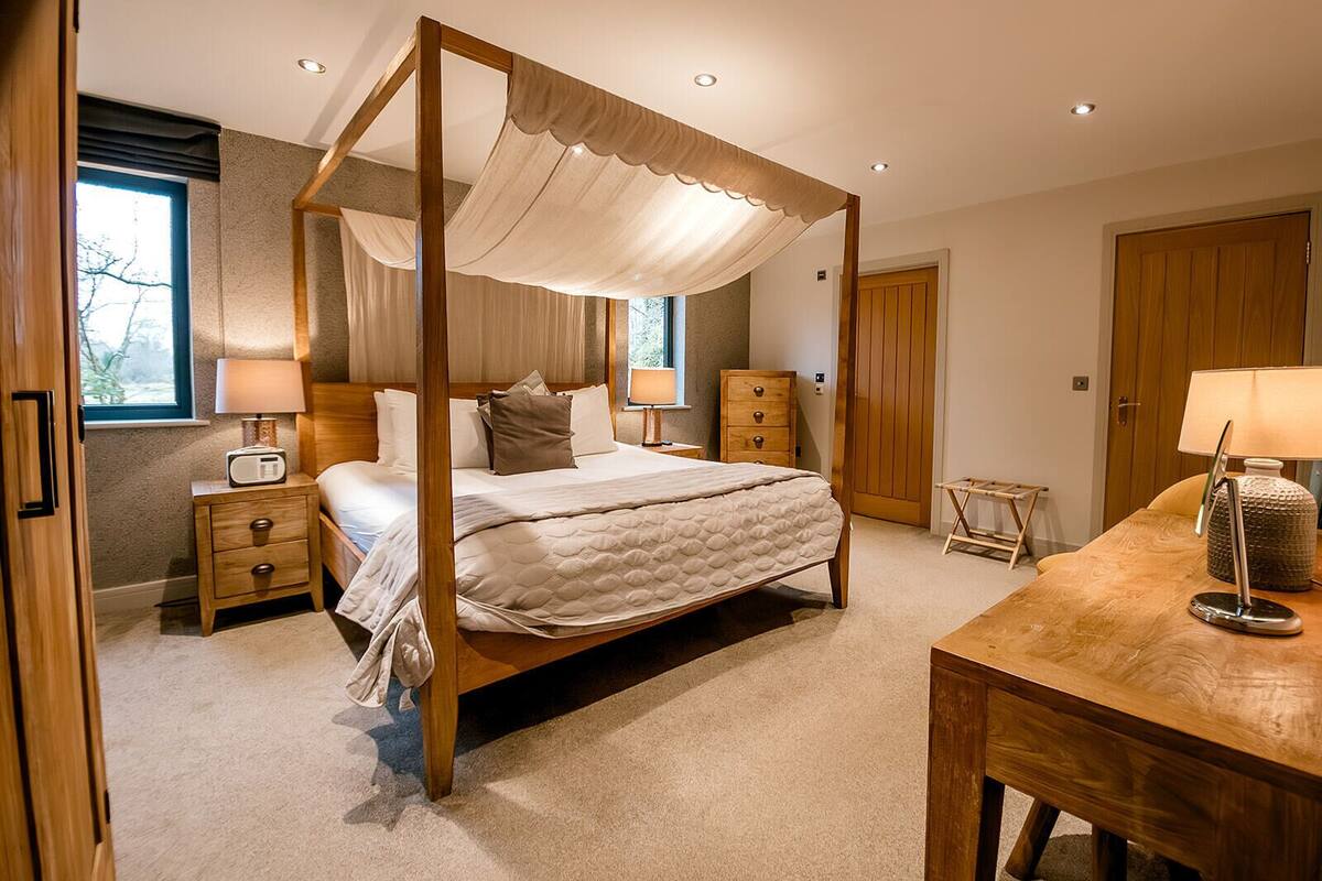 A four-poster canopy bed with a quilted mattress, bedside tables with lamps, a wooden dresser, and a wooden desk with a lamp.