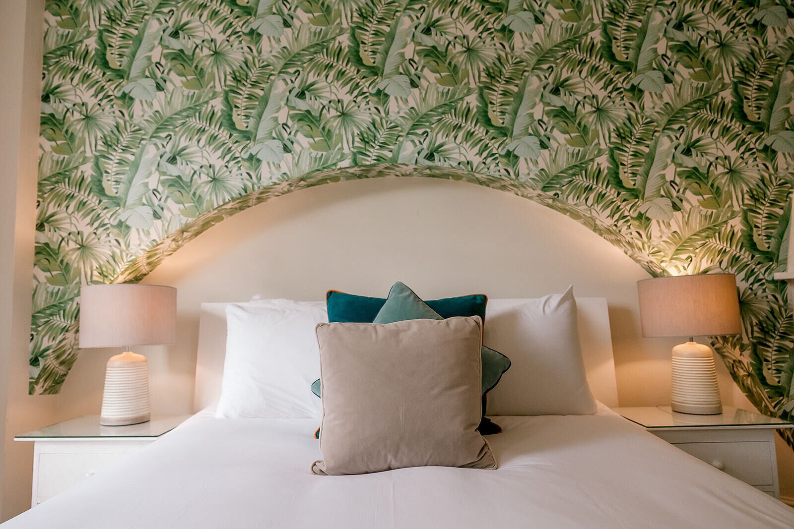 Een bed met wit beddengoed, kussens in teal en beige tinten, en twee bedlampjes.