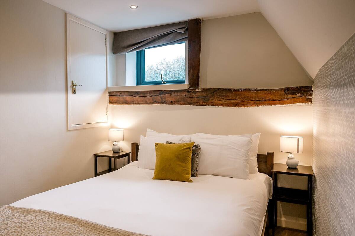 A bedroom with a large bed, two bedside tables with lamps, a window with a view, and exposed wooden beams.