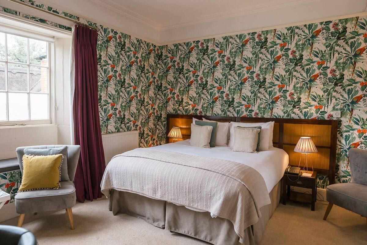 A bedroom with a floral wallpaper, a large bed with two bedside tables, a chair, and a window with curtains.