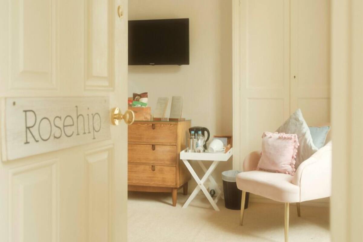 A room with a white door labeled "Rosehip," a wooden dresser, a small white table, a pink chair, and a television on the wall.