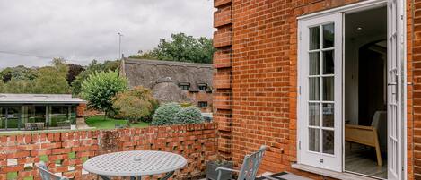 A patio with a round table and chairs, a brick wall, and a thatched-roof building in the background.