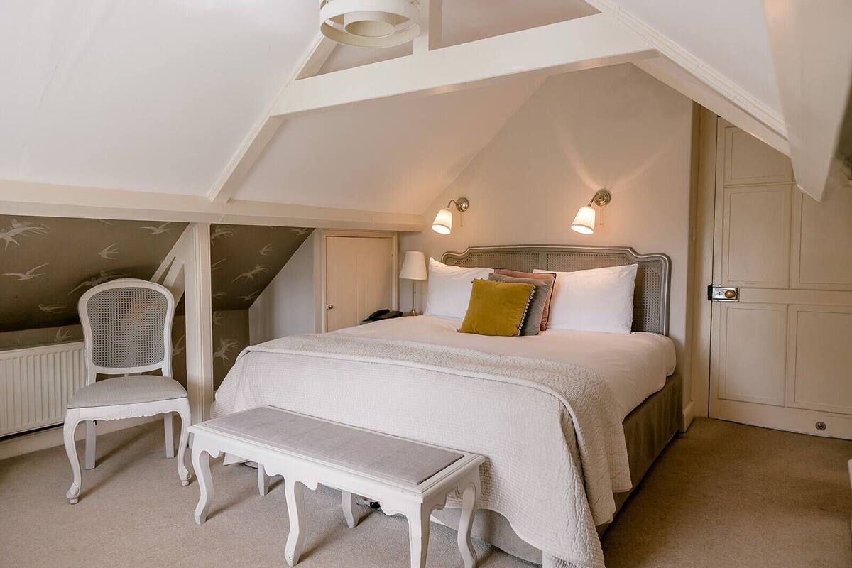 A bedroom with a sloped ceiling, a bed with white bedding, a bedside lamp, a chair, and a bench.