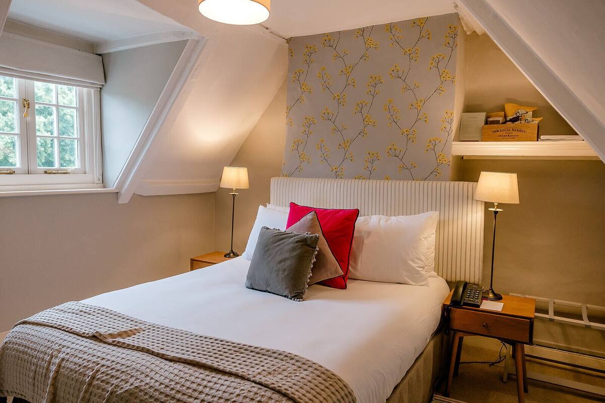 A bedroom with a bed, two bedside tables, a nightstand, a window with curtains, and a wall with a floral pattern.