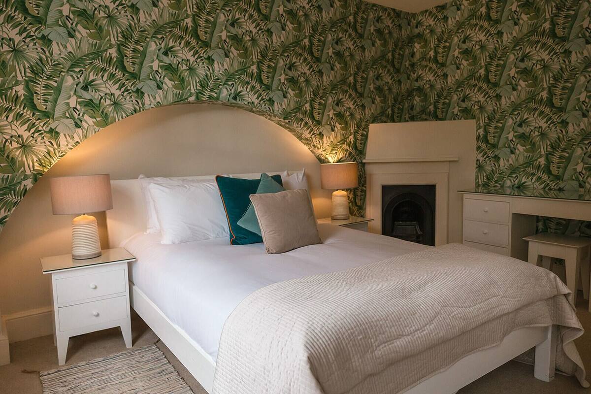 A bedroom with a bed, two bedside tables, a nightstand, a fireplace, and a desk.