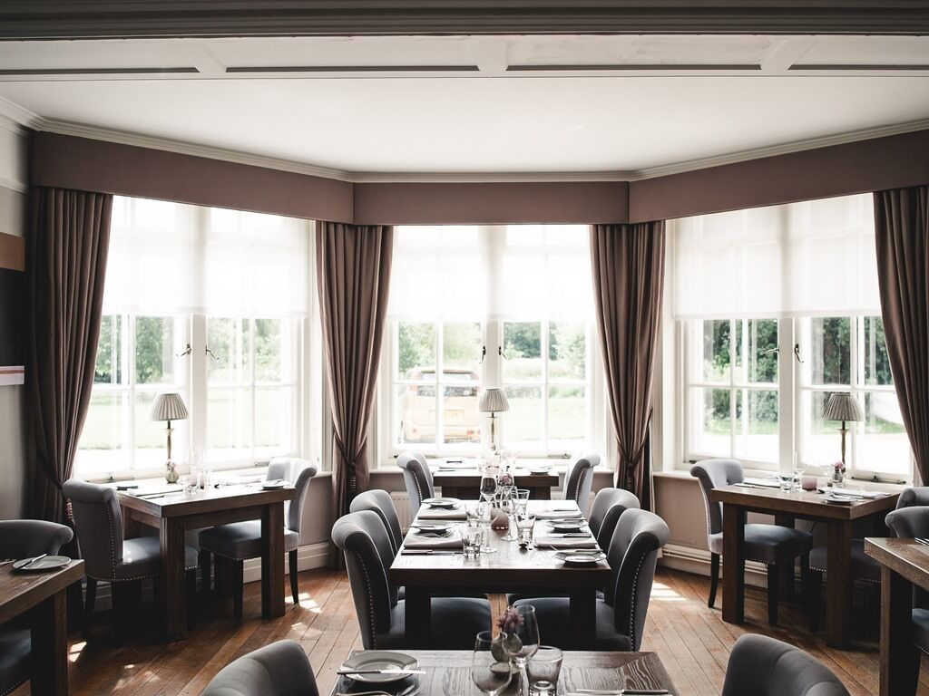 Un coin repas avec une longue table dressée pour six personnes, entourée de chaises, et de grandes fenêtres agrémentées de rideaux.