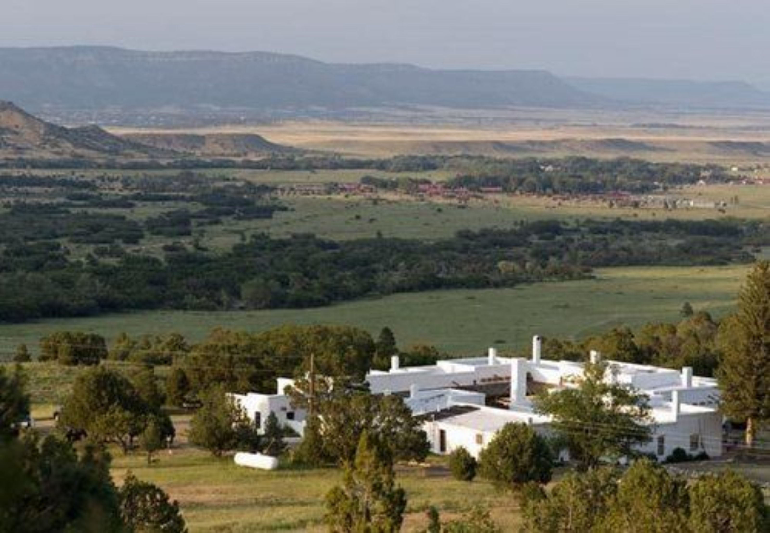 A white building with multiple chimneys, surrounded by trees and open fields, with distant hills in the background.