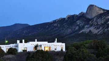 Un edificio blanco con numerosas ventanas y una puerta, rodeado de follaje y montañas al fondo.