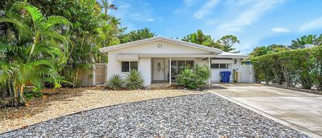A single-story house with a front porch, surrounded by tropical vegetation and a gravel driveway.