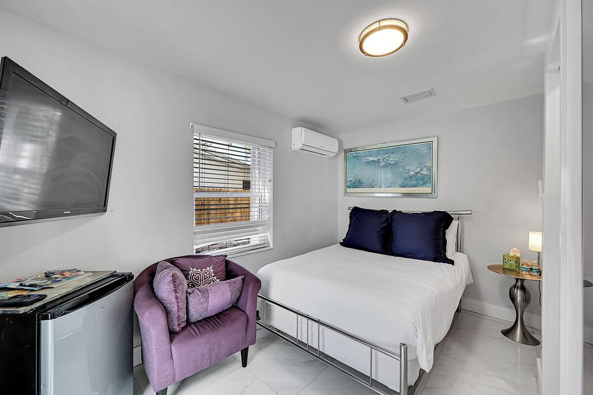 A bedroom with a bed, a TV, a window with blinds, an air conditioner, a painting, and a small table with a lamp.