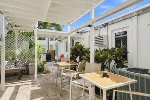 Un espace extérieur aménagé avec des tables et des chaises, protégé par un auvent, et entouré de plantes en pot et d’un bâtiment blanc.