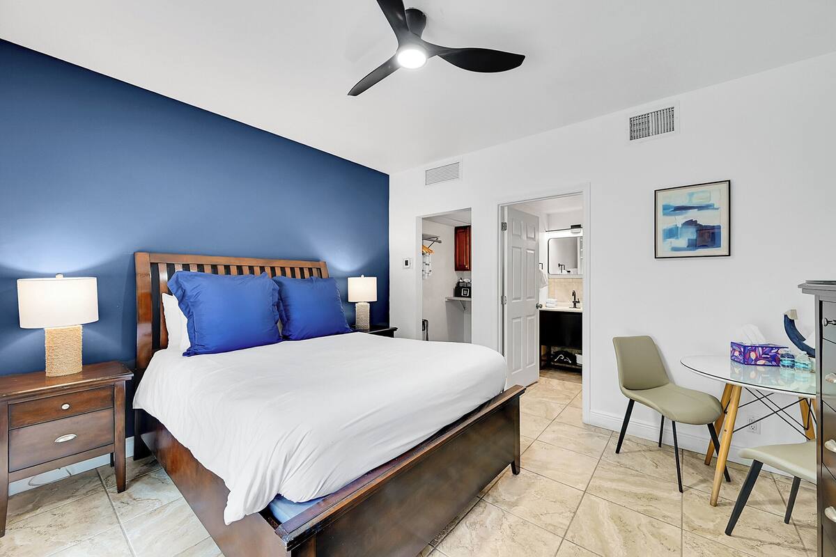 A bedroom with a bed, bedside tables, a ceiling fan, a desk with a chair, and a painting on the wall.