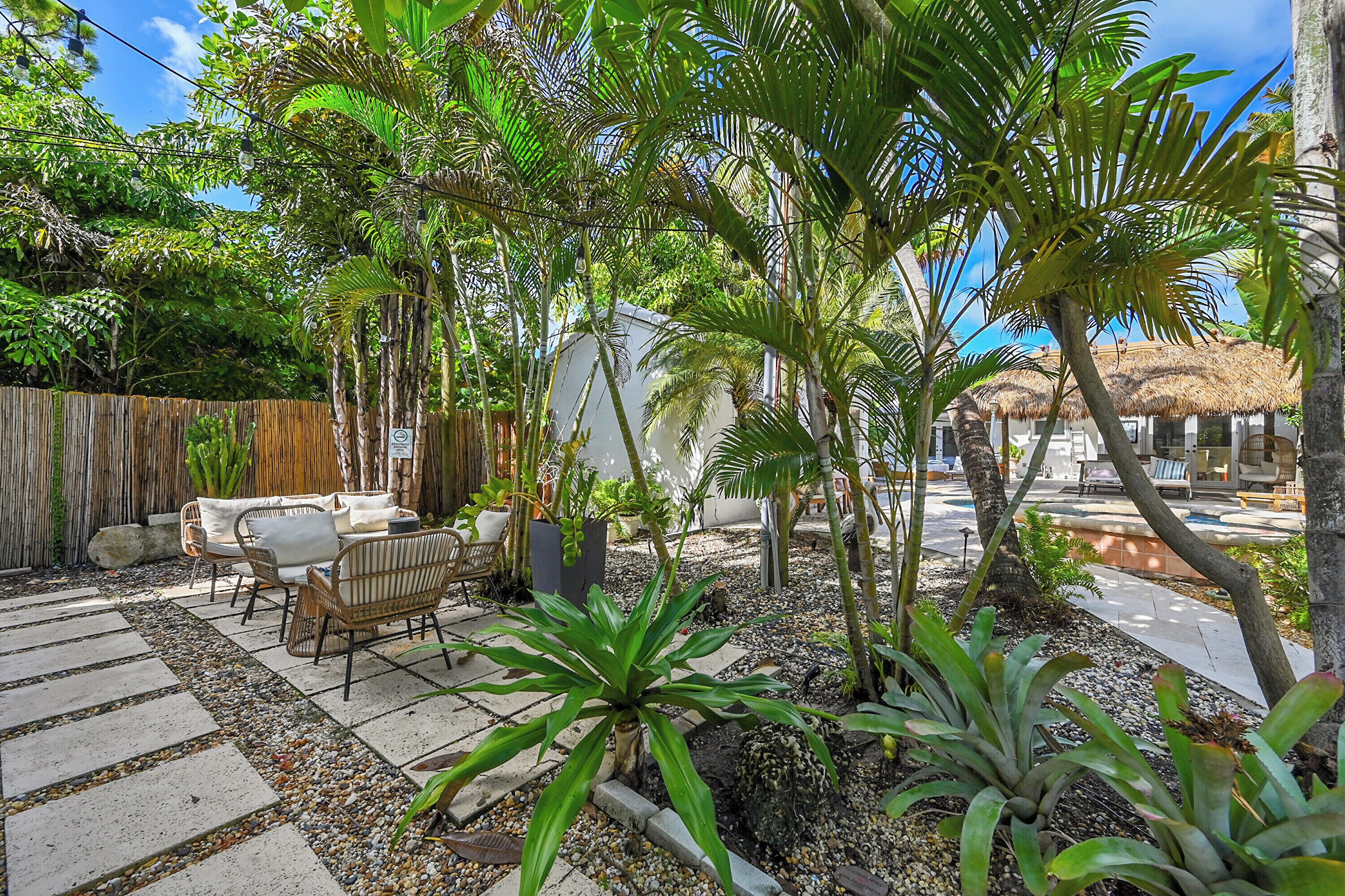 Um pátio com mesa e cadeiras sob um telhado de palha, cercado por plantas tropicais e uma cerca de madeira.