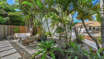 Um pátio com mesa e cadeiras sob um telhado de palha, cercado por plantas tropicais e uma cerca de madeira.