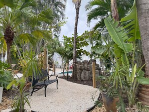 Un espace jardin agrémenté d’un banc, de différentes plantes, d’un petit bassin et d’un bâtiment blanc en arrière-plan.