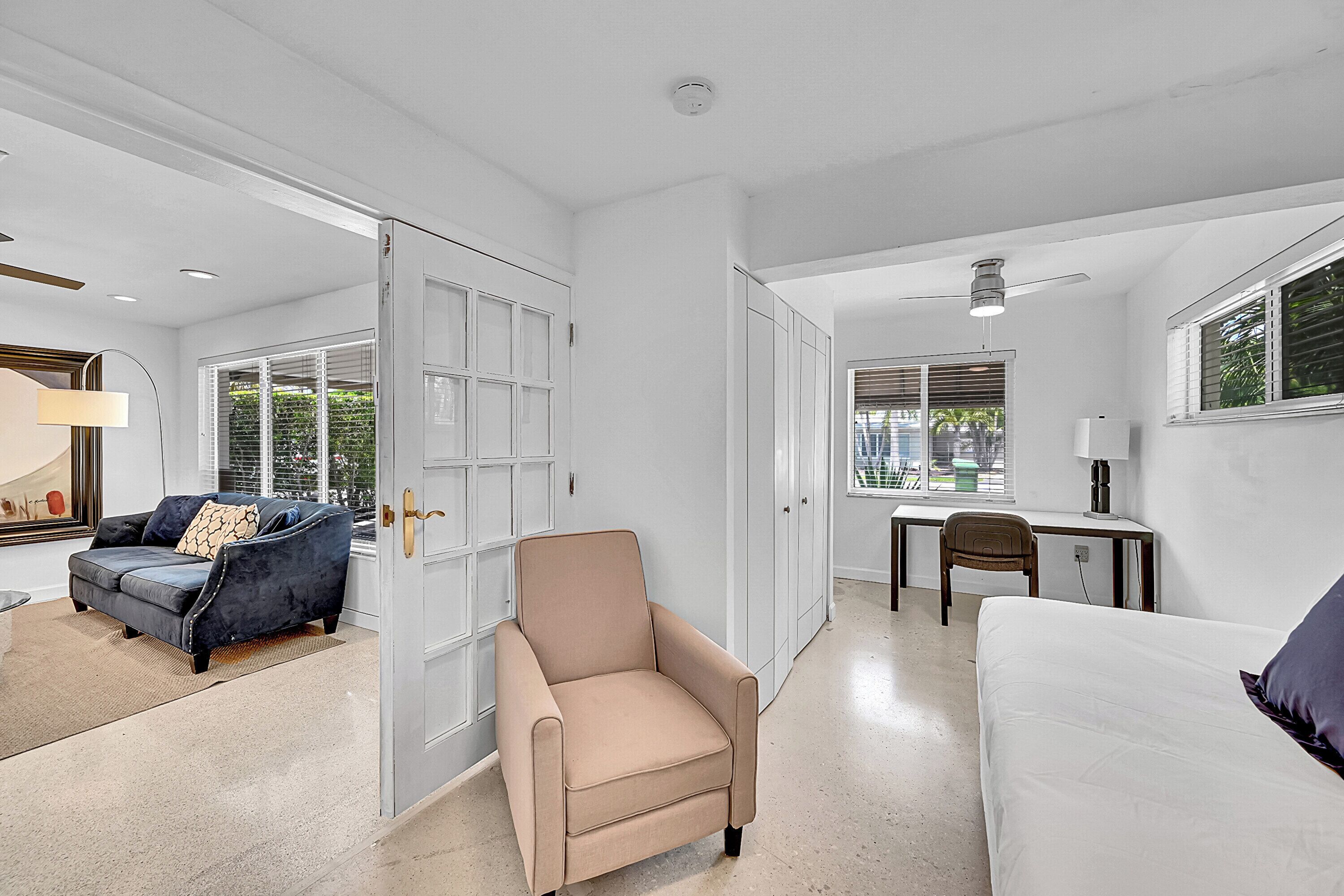 Une pièce spacieuse avec un fauteuil beige, un canapé blanc, un bureau avec une chaise et une grande fenêtre équipée de stores.