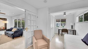 Une pièce spacieuse avec un fauteuil beige, un canapé blanc, un bureau avec une chaise et une grande fenêtre équipée de stores.