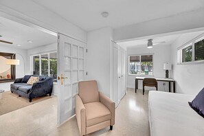 Ein geräumiger Raum mit einem beigefarbenen Sessel, einem weißen Sofa, einem Schreibtisch mit Stuhl und einem großen Fenster mit Jalousien.