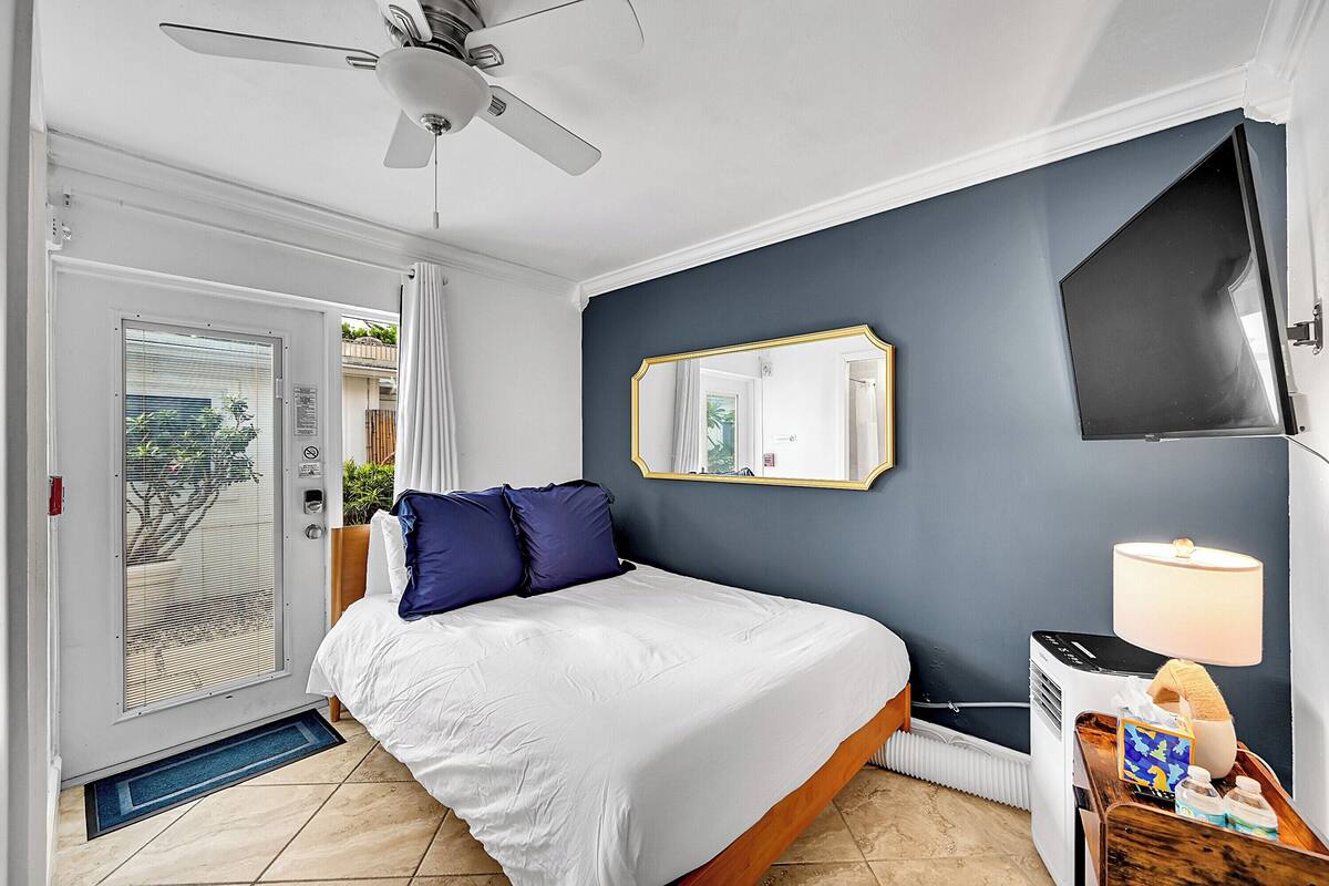 A hotel room with a bed, a ceiling fan, a TV, and a bedside table with a lamp and a tray with toiletries.