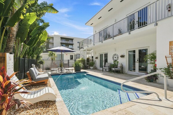 Une piscine avec des chaises longues, un espace de détente couvert et un bâtiment doté de balcons.