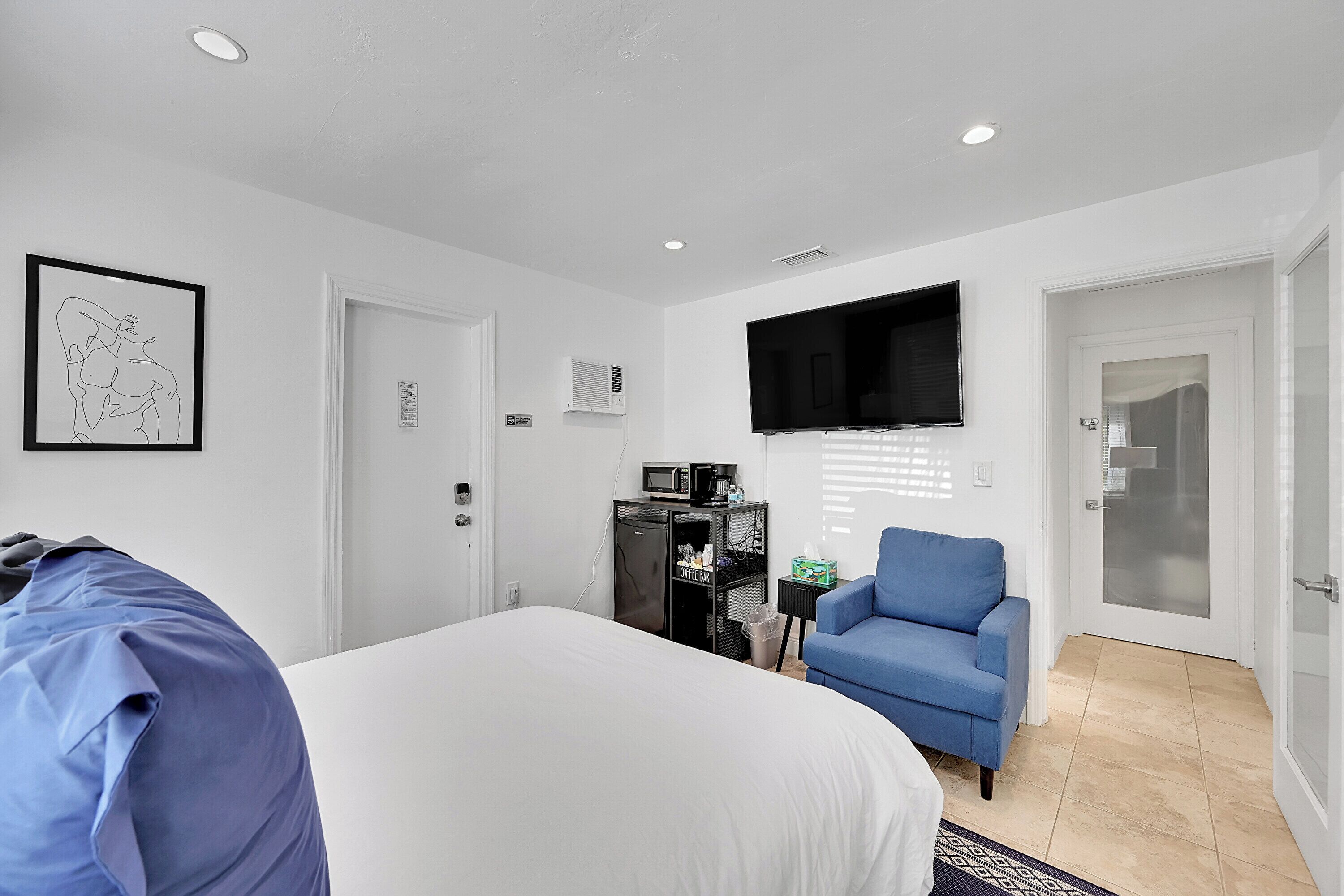 A hotel room with a bed, a blue armchair, a TV mounted on the wall, a coffee machine, and a door leading to another room.