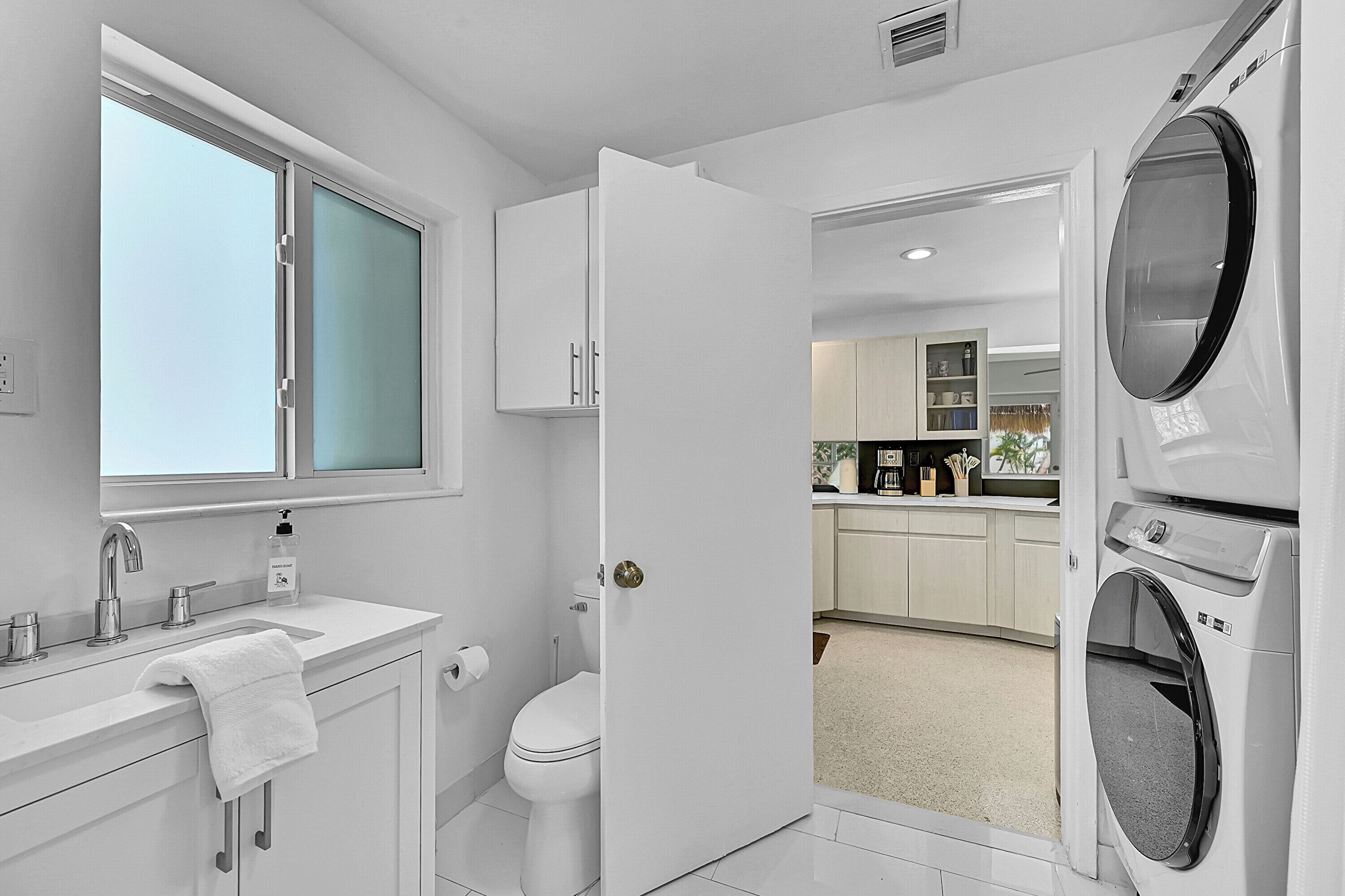 A modern bathroom with a sink, toilet, and open door leading to a laundry area with a washing machine.