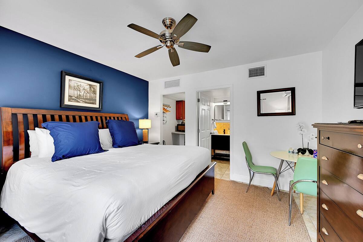 A hotel room with a large bed, a ceiling fan, a desk with a chair, and a dresser.