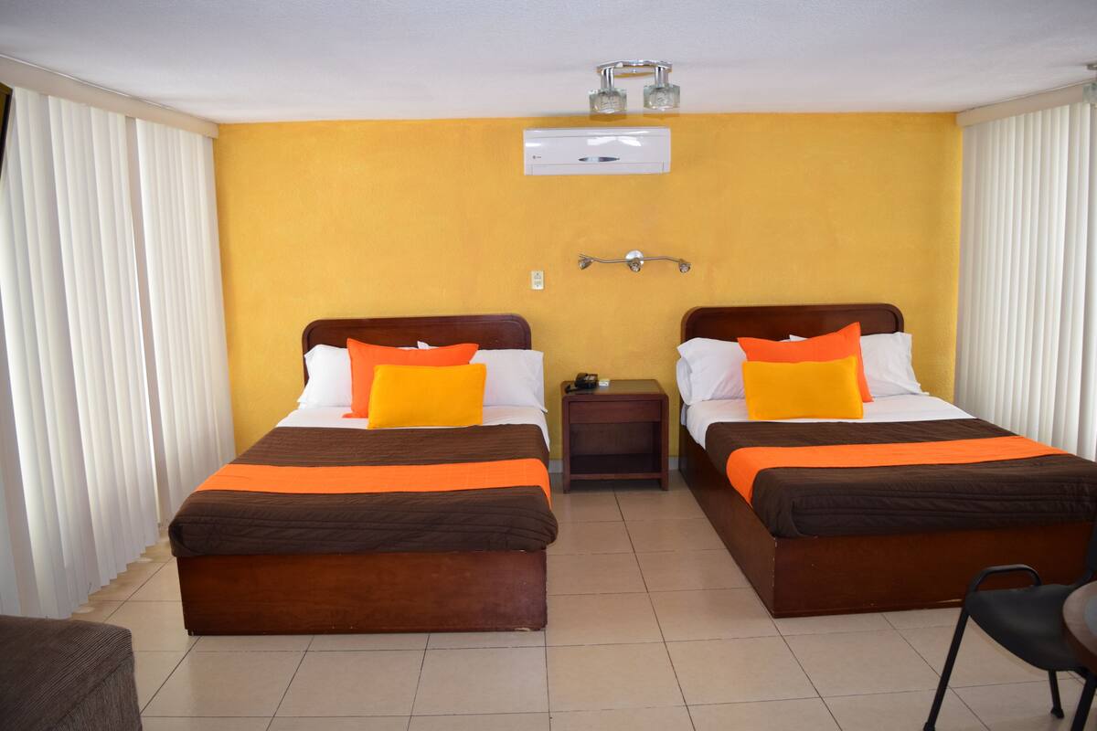 A hotel room with two beds, each with a brown headboard, white linens, and orange pillows. There is a small bedside table with a lamp between the beds, and a window with white blinds.