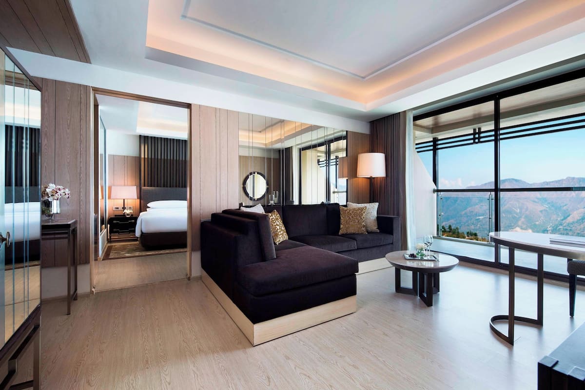 Une chambre d’hôtel moderne avec un grand lit, un canapé de couleur sombre, une petite table ronde et une grande fenêtre donnant sur une vue imprenable sur les montagnes.