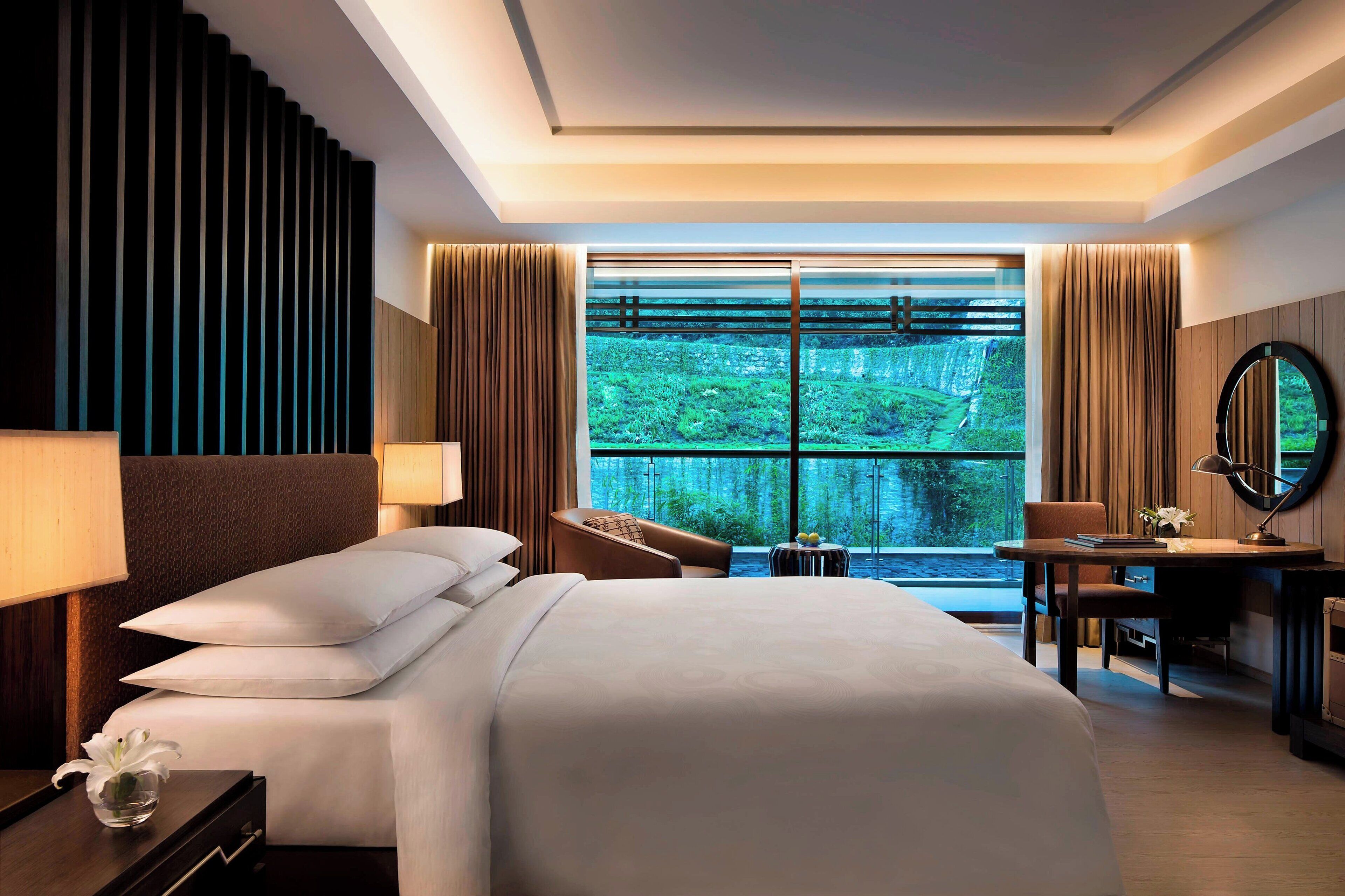A modern hotel room with a large bed, a desk, and a view of a lush garden through floor-to-ceiling windows.
