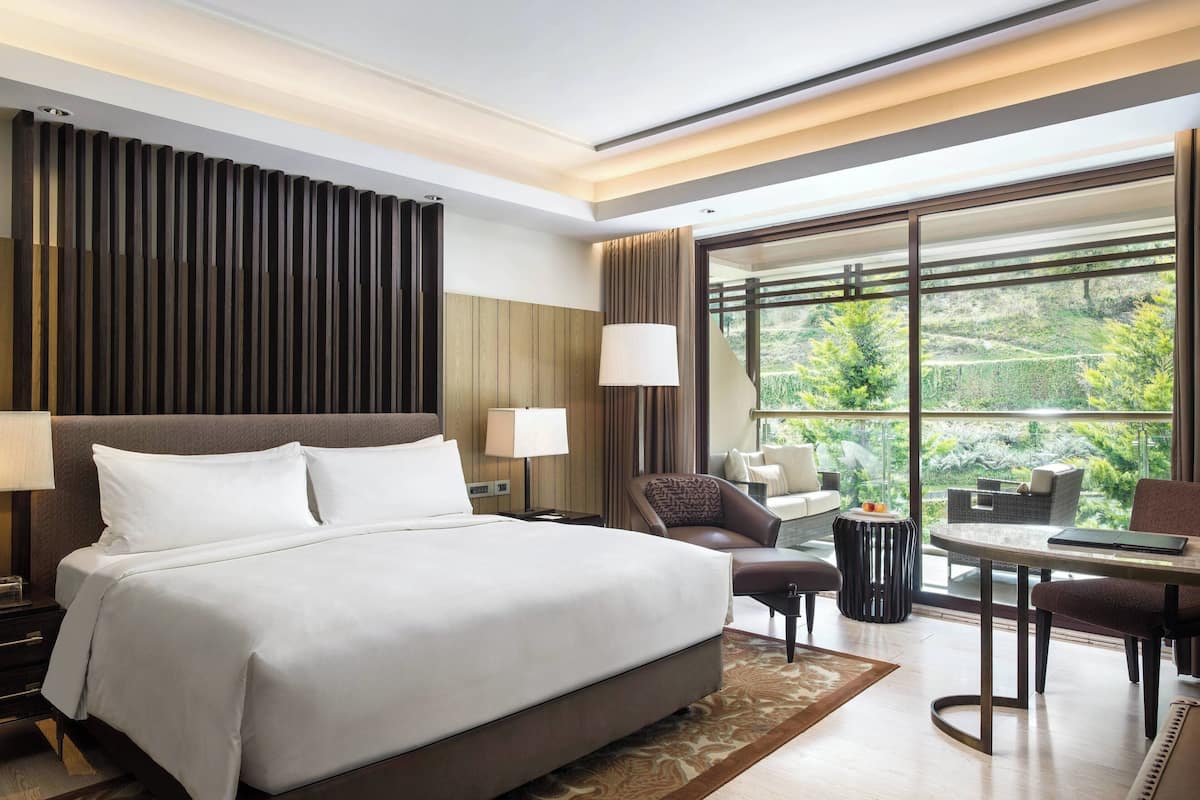 Une chambre d’hôtel moderne dotée d’un grand lit, d’un coin salon avec une table et des chaises, et offrant une vue sur la verdure grâce à de grandes fenêtres.