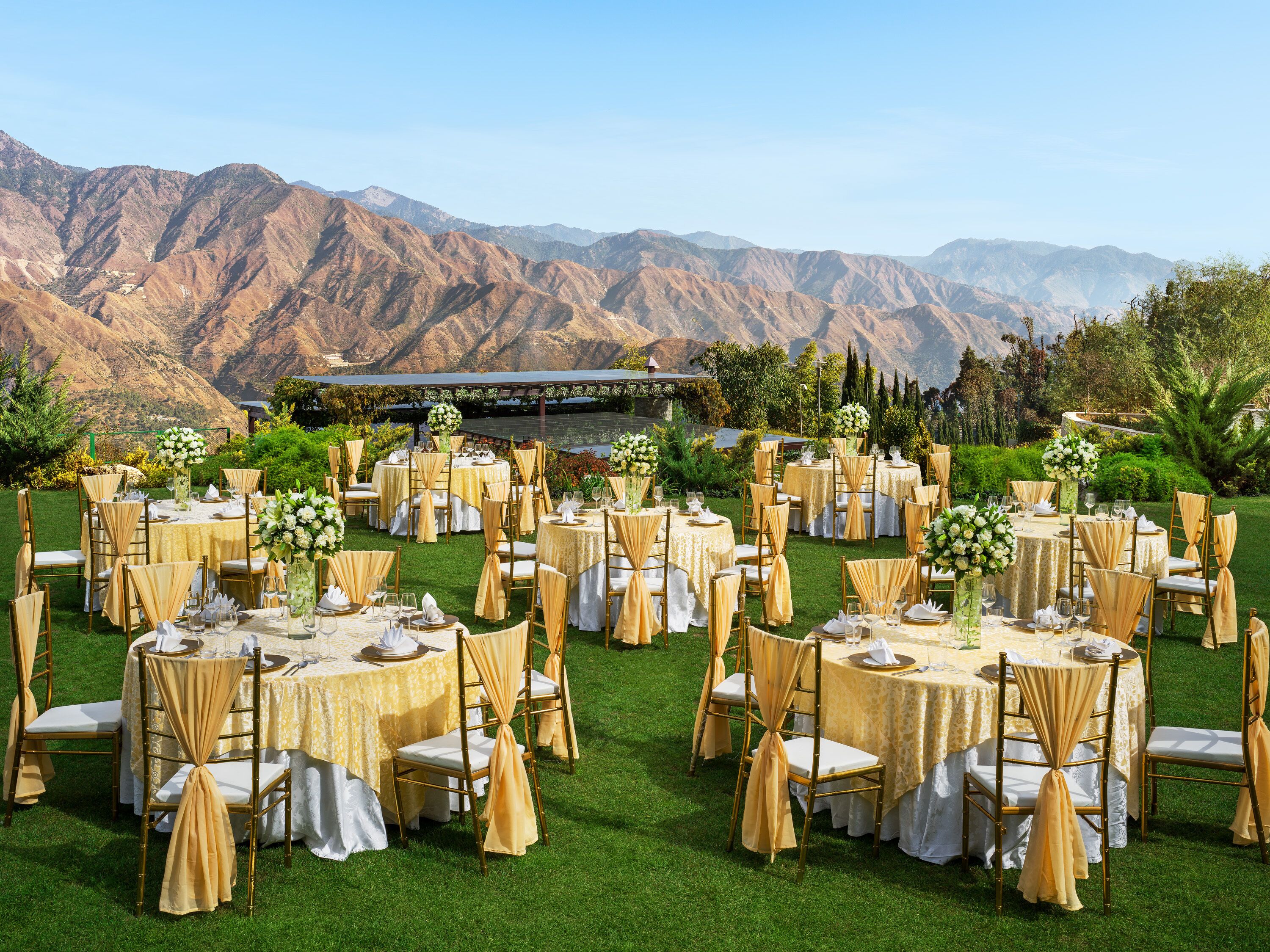 An outdoor wedding venue with round tables, gold chairs, and white tablecloths set against a backdrop of mountains.