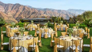 An outdoor wedding venue with round tables, gold chairs, and white tablecloths set against a backdrop of mountains.