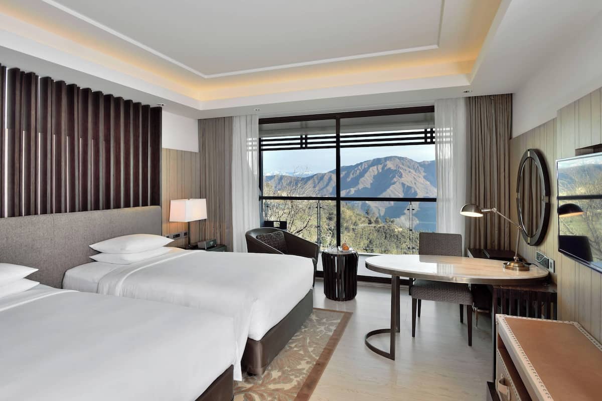 Une chambre d’hôtel avec deux lits, un bureau, une chaise, une télévision et une grande fenêtre donnant sur la montagne.
